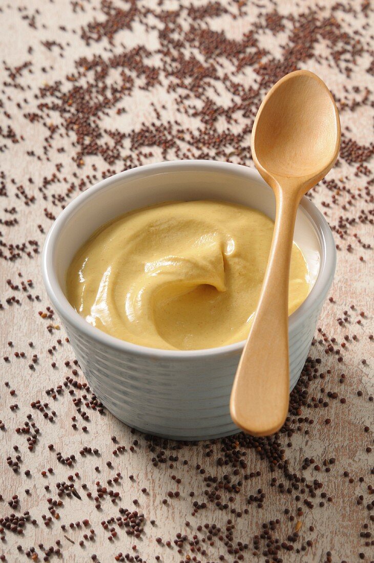 A bowl of mustard with a spoon surrounded by mustard seeds