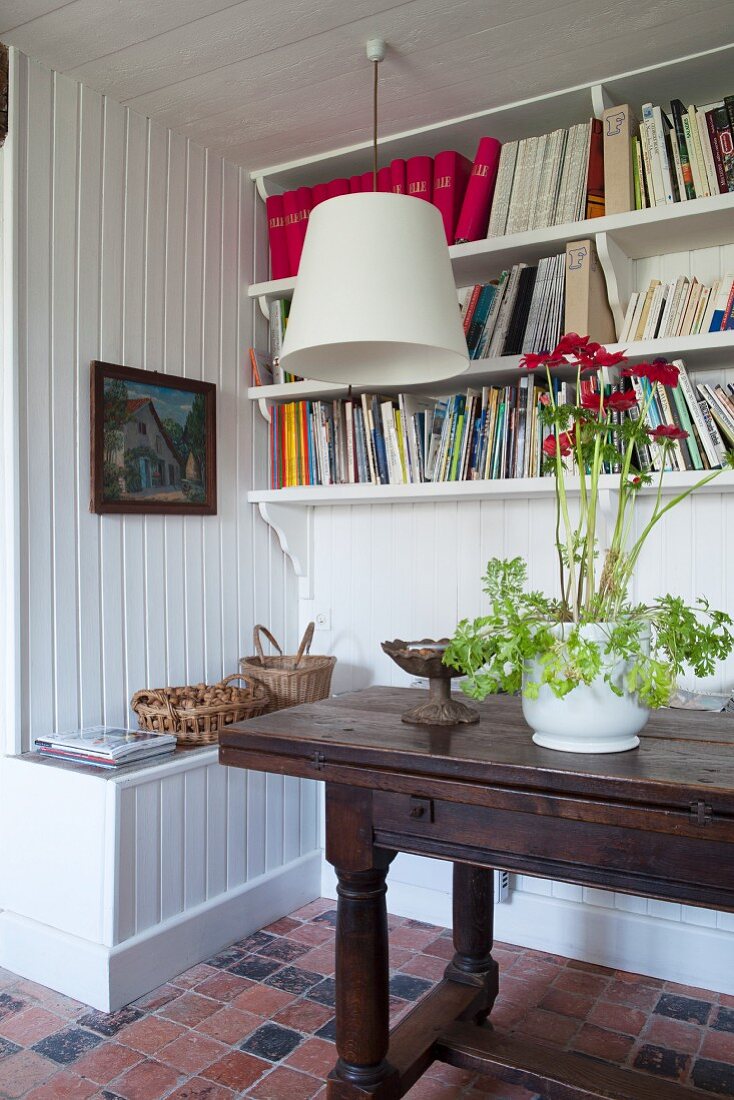 Flowering potted plant on antique console table below bookshelves on white wooden wall