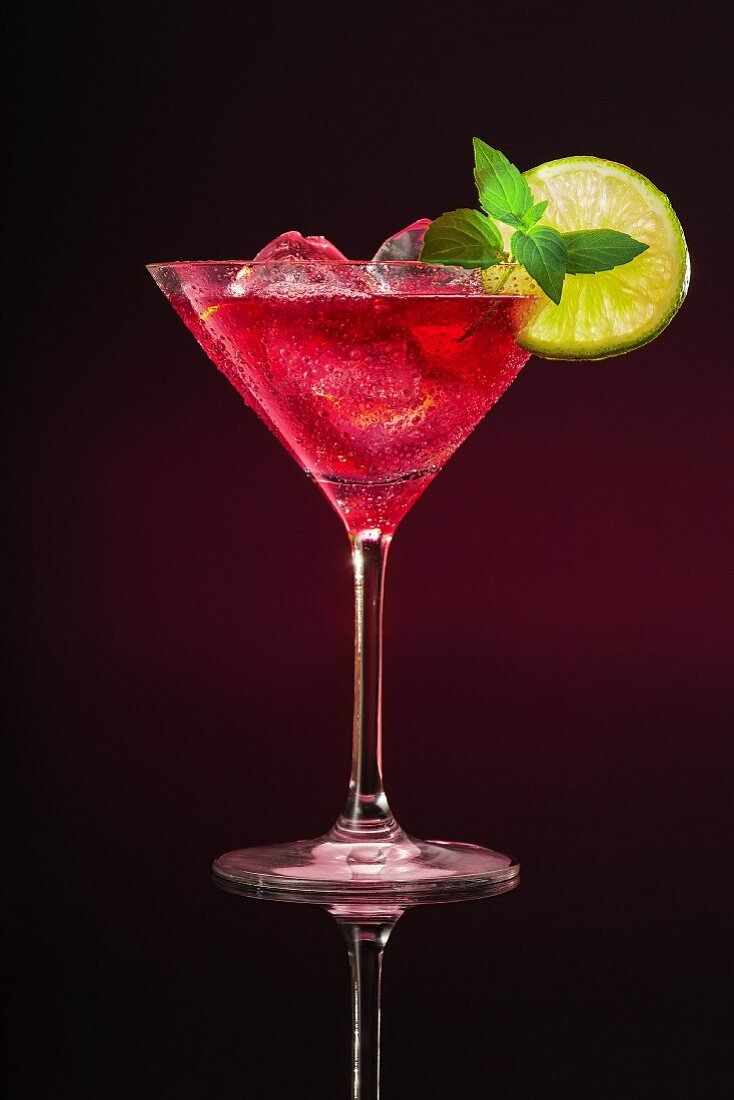 Aping cocktail in a glass garnished with a lime slice and a sprig of mint against a dark background