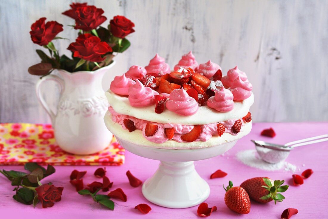 Meringue cake with cream, strawberries and rose petals on a cake stand in front of a vase of red roses