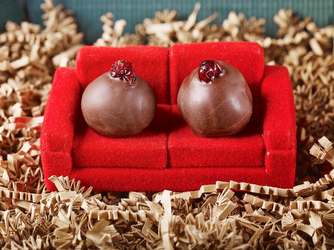 Zwei handgerollte Marzipantrüffeln in Zartbitterkuvertüre mit Cranberries als Couchpotato auf einem roten Sofa