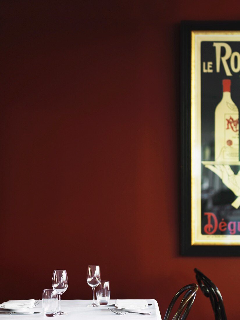 A laid table with wine and water glasses against a dark red wall with a retro advertising poster
