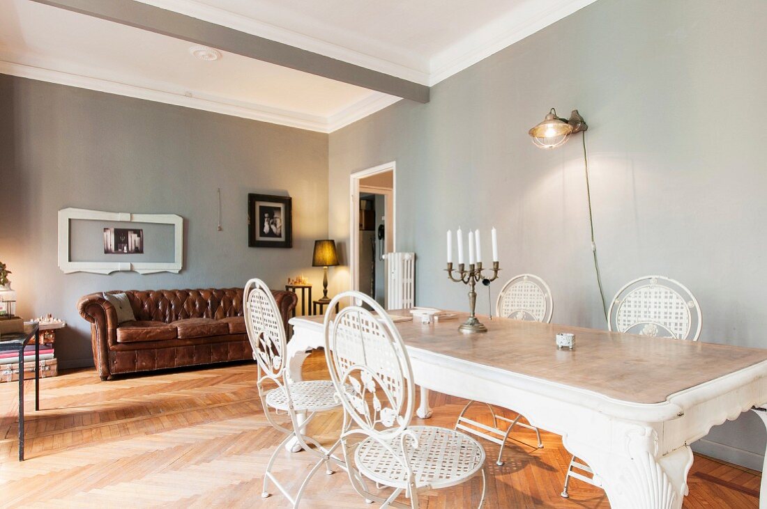 White metal chairs around dining table in front of brown leather couch in room with walls painted light grey