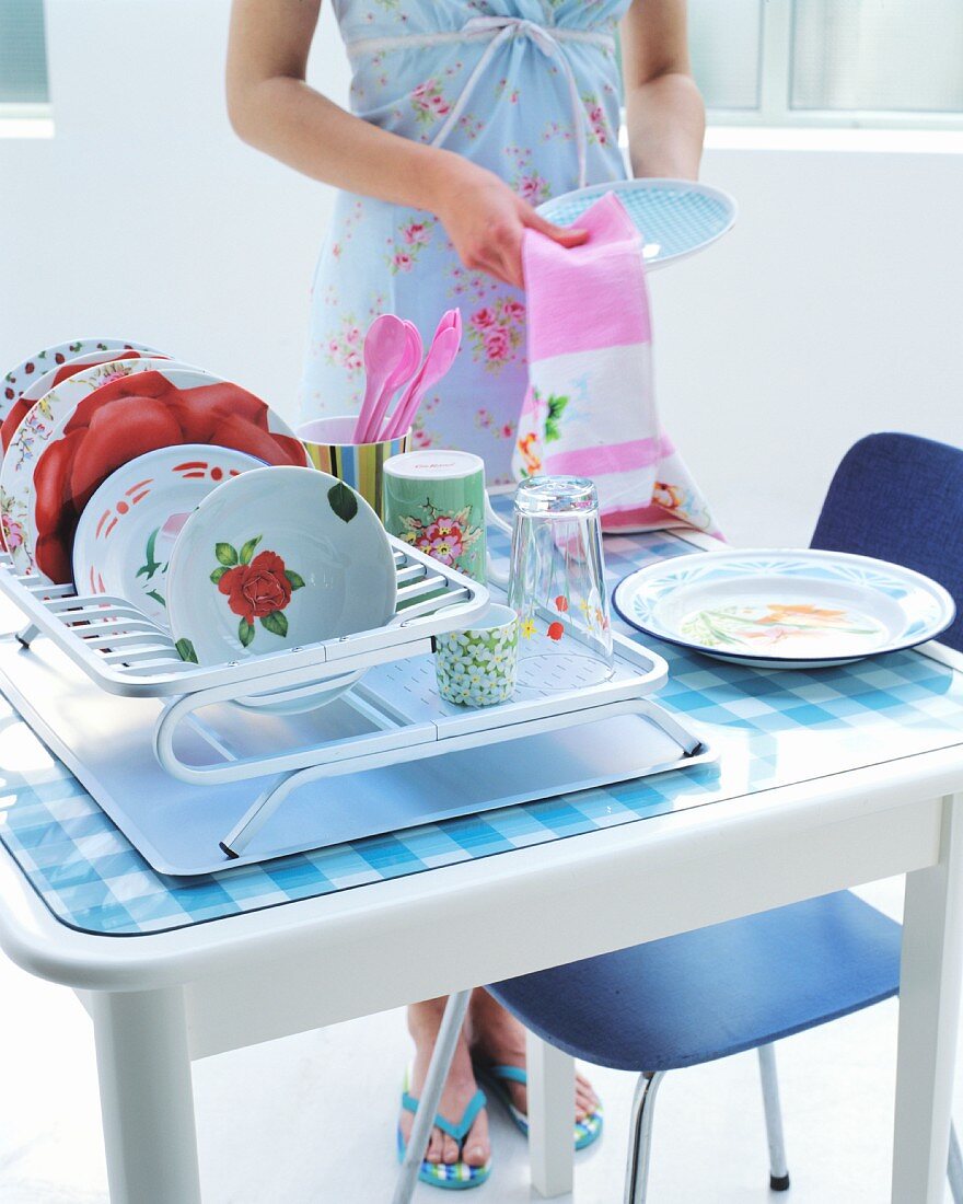 Floral crockery in drying rack on kitchen table; woman holding tea towel and pale blue plate