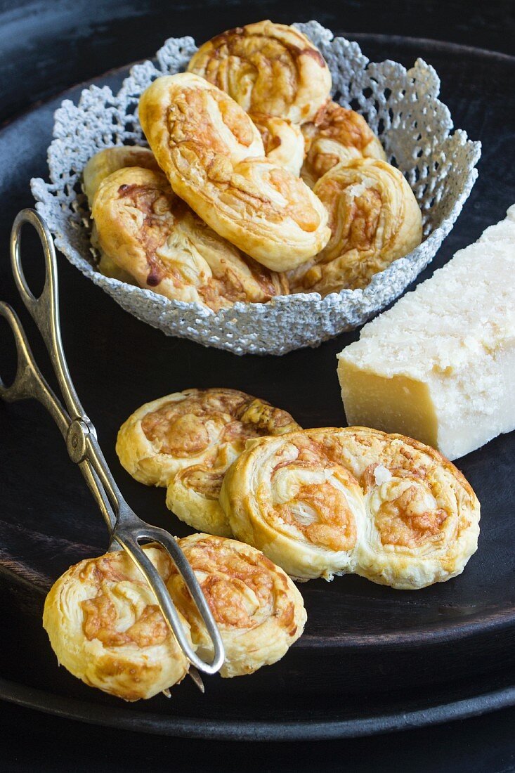 Savoury puff pastries with Parmesan cheese on a black plate with a pair of pastry tongs