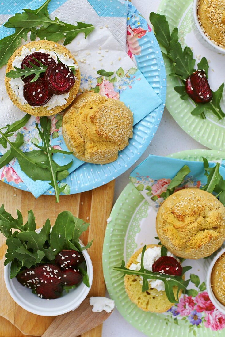 Glutenfreie Sesambrötchen auf bunten Papptellern, serviert mit roten Rüben und Rucola