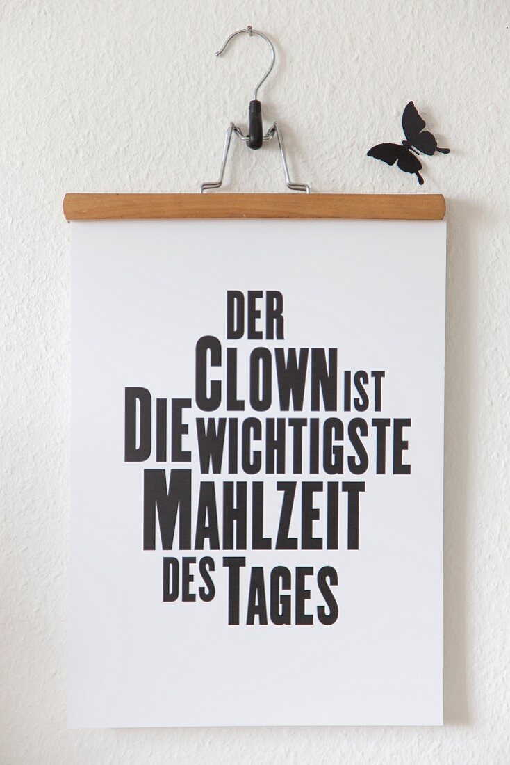 Aufgehängter Hosenbügel mit Sinnspruch und schwarzem Dekoschmettterling an weißer Wand