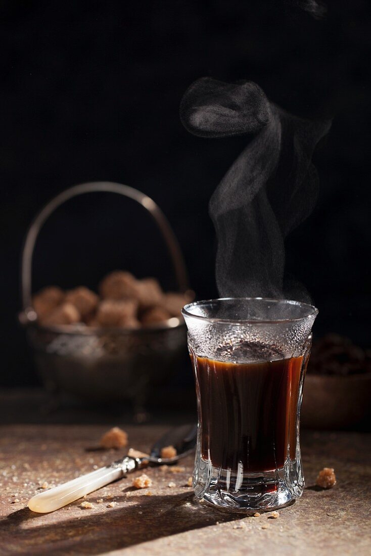 A glass of steaming black coffee with a bowl of sugar cubes in the background