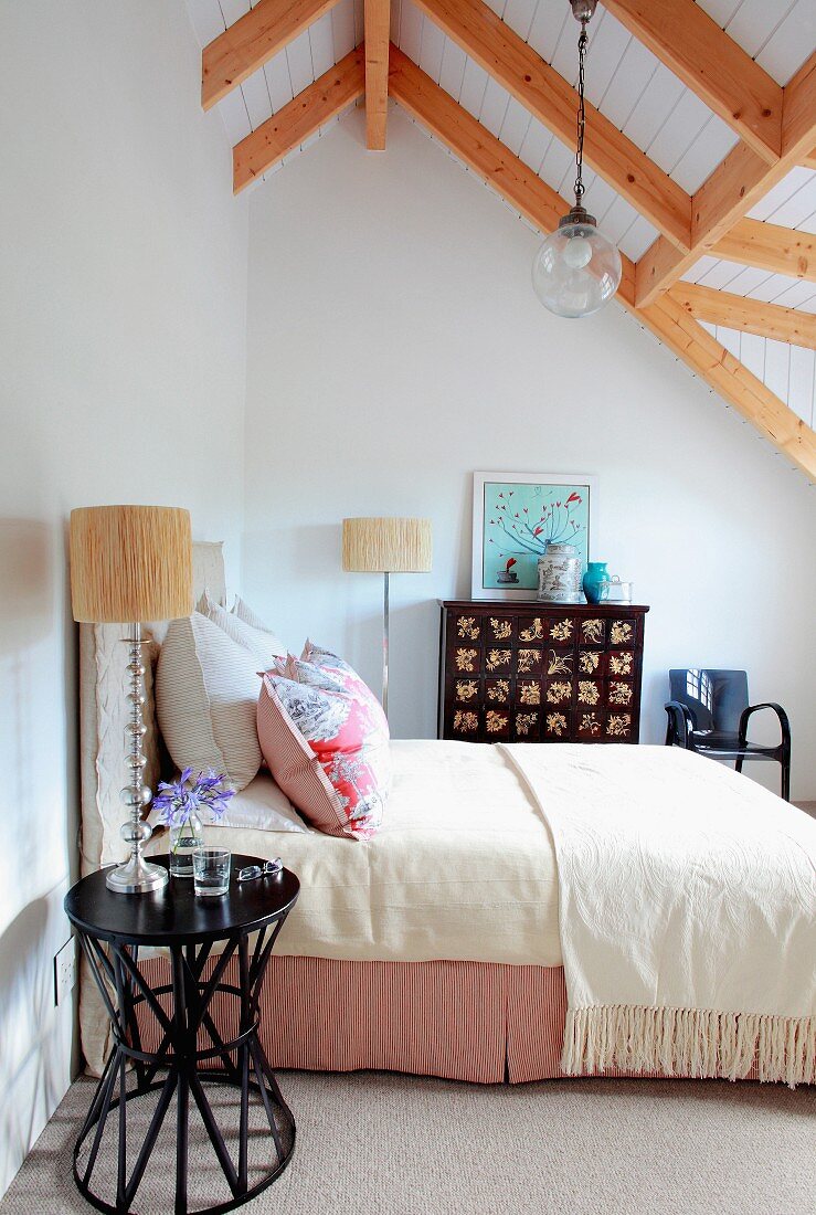 Black side table next to double bed with pale bedspread in attic room with exposed wooden ceiling beams
