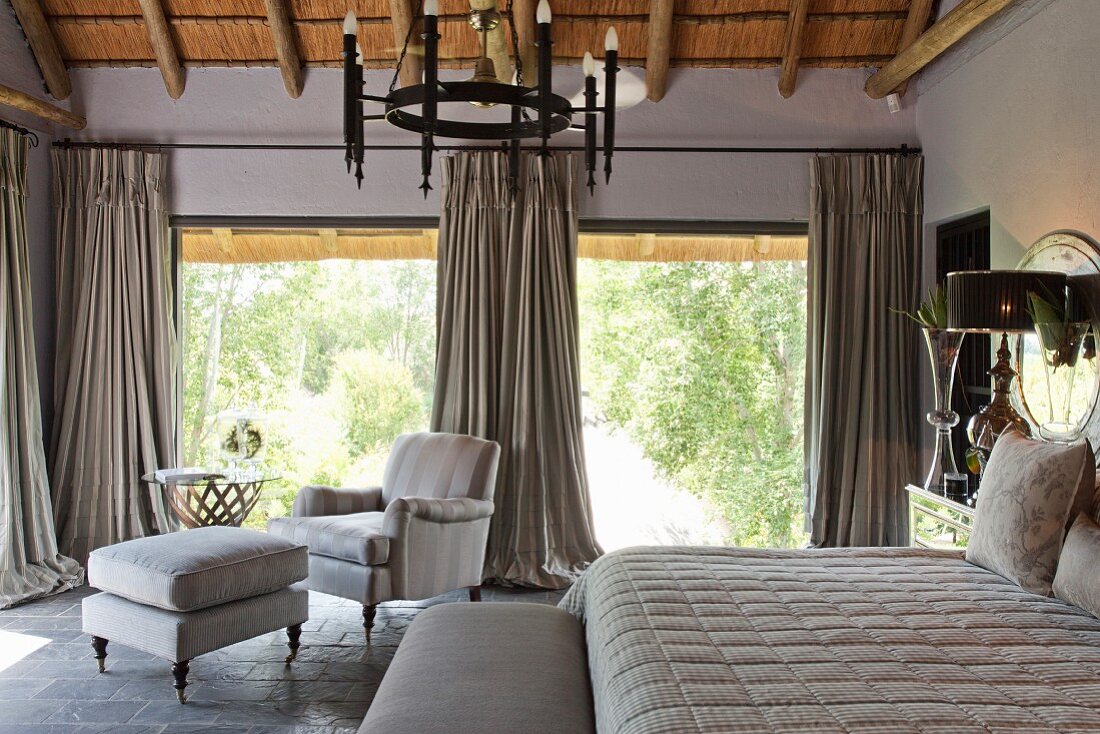 Doppelbett und Polstersessel mit passendem Fußschemel vor offenen Terrassentüren, bodenlange Vorhängen und Gartenblick im eleganten Vintage Schlafzimmer