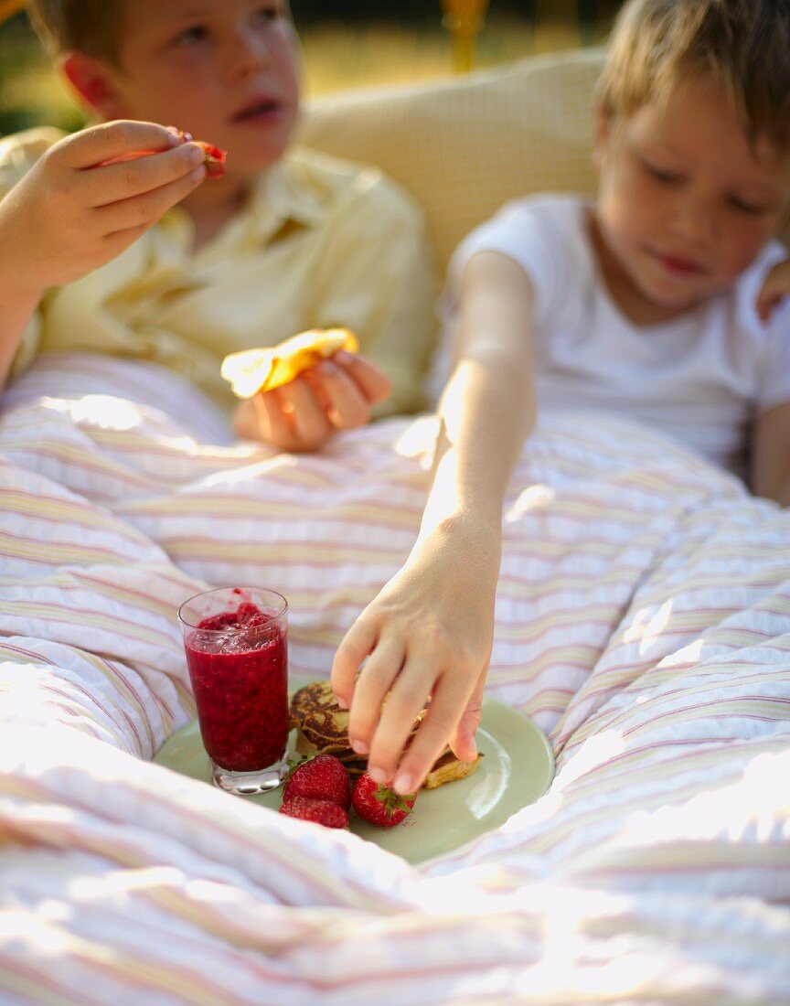 Two boys eating pancakes with strawberries for breakfast in bed in a field