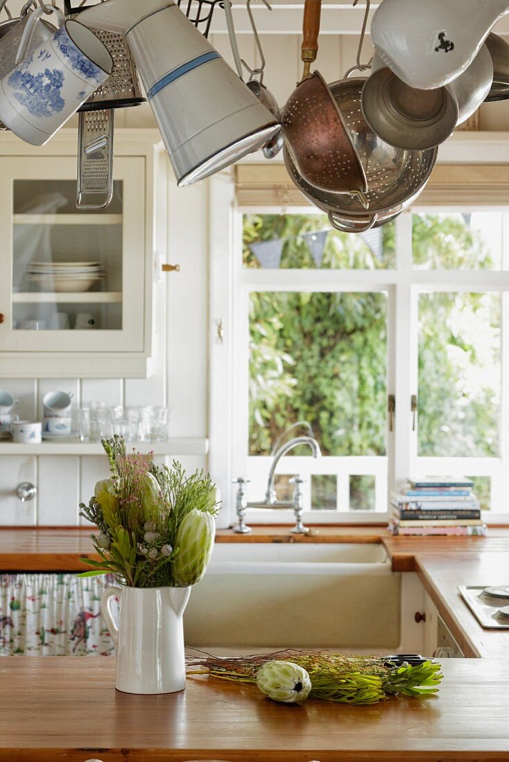 Tropical plants in white china jug on kitchen counter below utensils suspended from rack in rustic kitchen