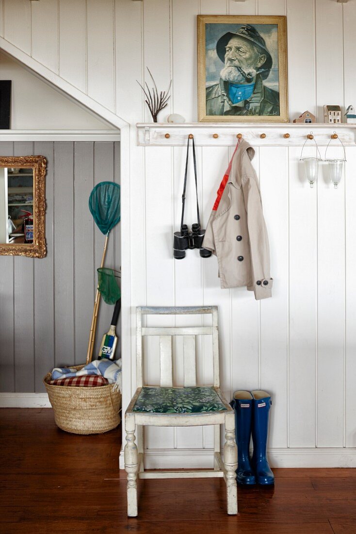 Kitchen chair below portrait and coat pegs on white, wood-panelled wall in foyer