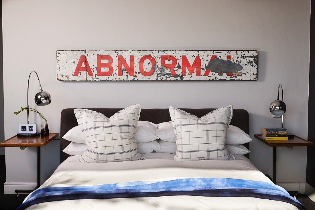 Double bed with checked pillows flanked by retro table lamps on bedside tables below vintage wooden sign with lettering