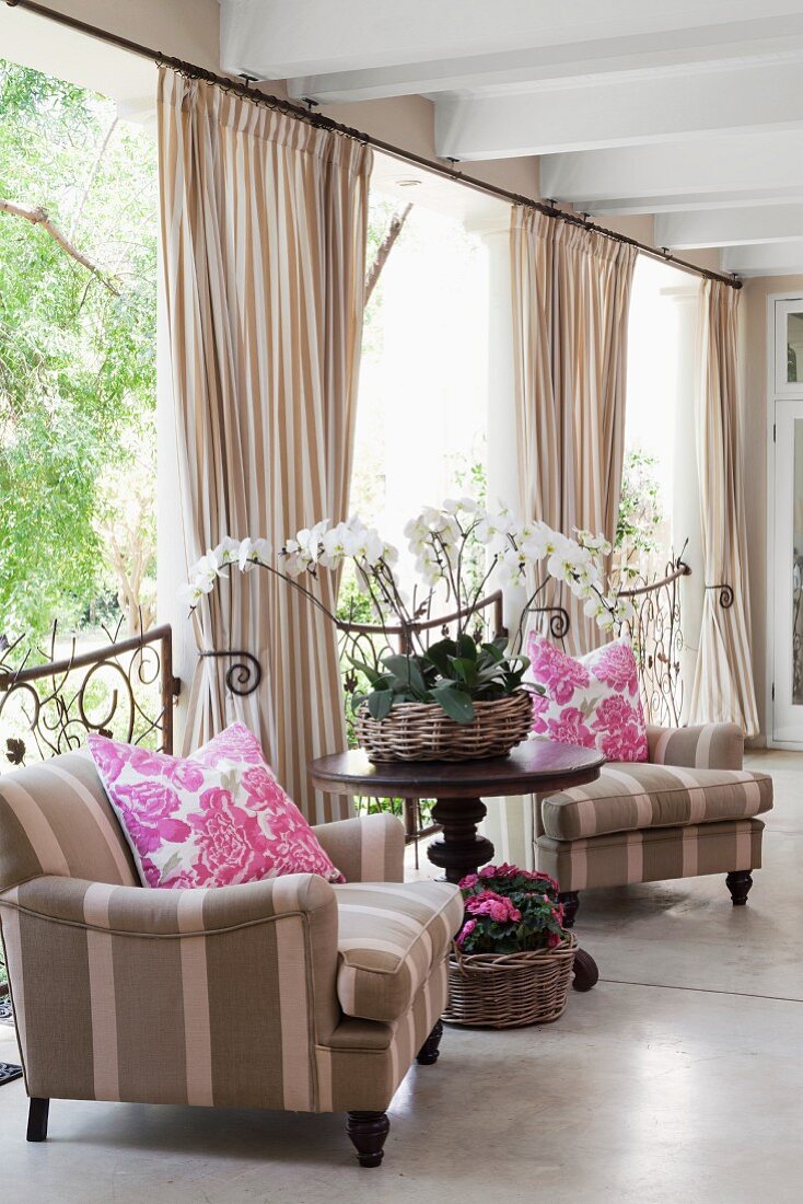 Armchairs with striped upholstery in grey and white either side of round table in front of gathered curtains and half-height grilles leading to veranda
