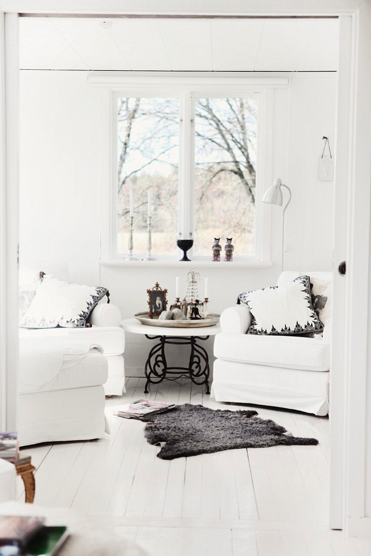 View through wide doorway with open sliding door, wooden floor and grey animal-skin rug in front of white armchairs below window