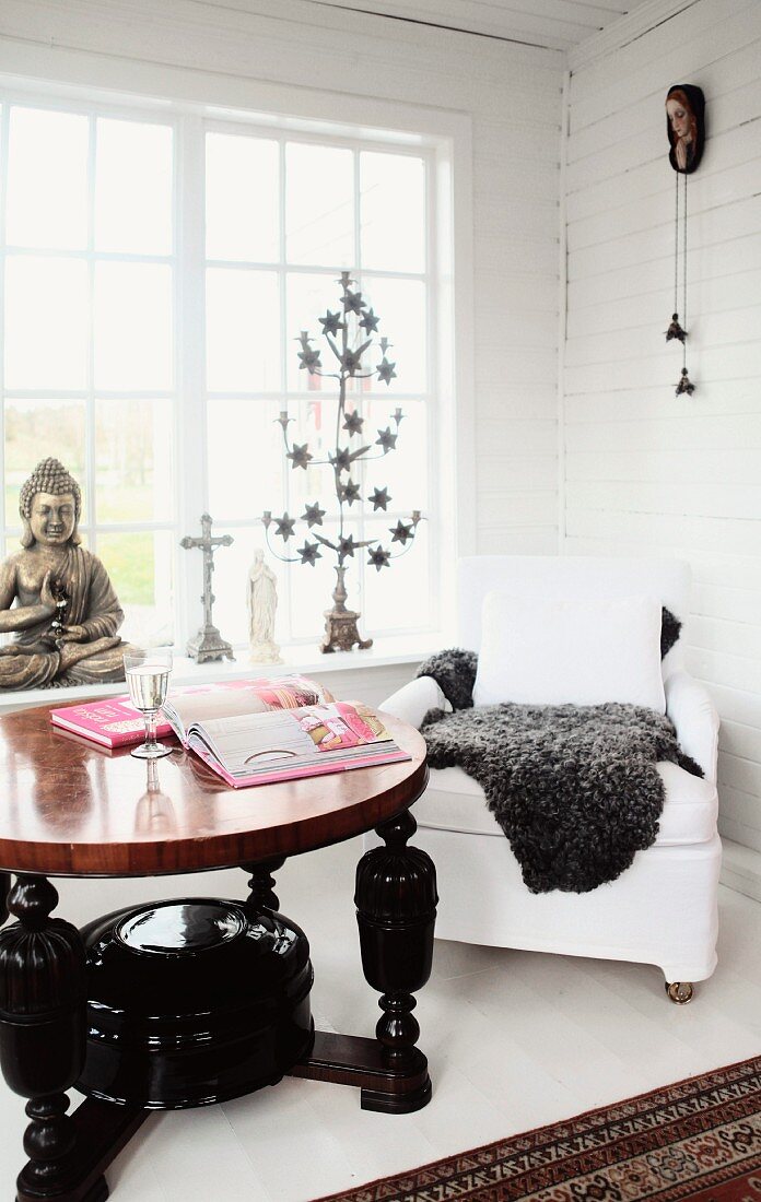 Runder Holztisch mit gedrechselten Beinen, schwarz lackiert und weisser Polstersessel vor Sprossenfenster in holzverkleideter Zimmerecke