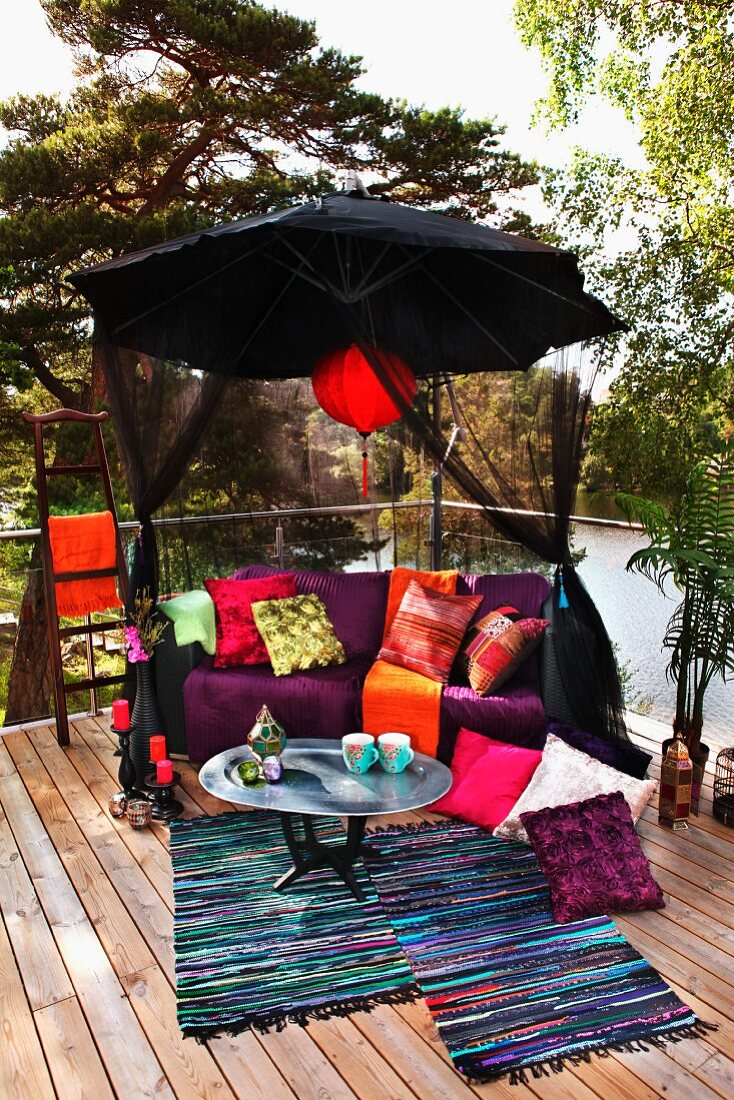 Terrassenecke mit farbenfrohem Ruheplätzchen unter schwarzem Sonnenschirm mit rotem Lampion, Seeblick mit Bäumen