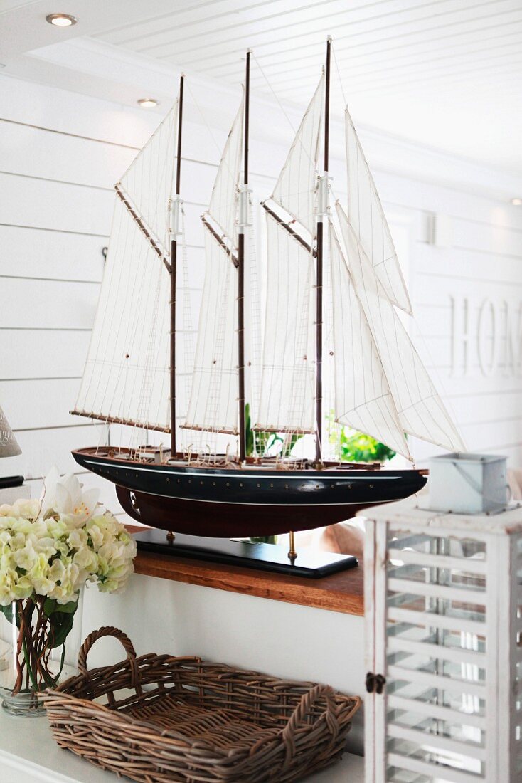 Splendid model of sailing boat on half-height wall in Scandinavian interior; wicker tray and vintage lantern in foreground