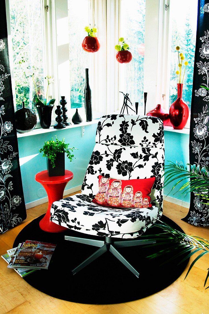 Swivel easy chair with black and white floral upholstery on round rug in corner; collection and black and red vases on windowsill