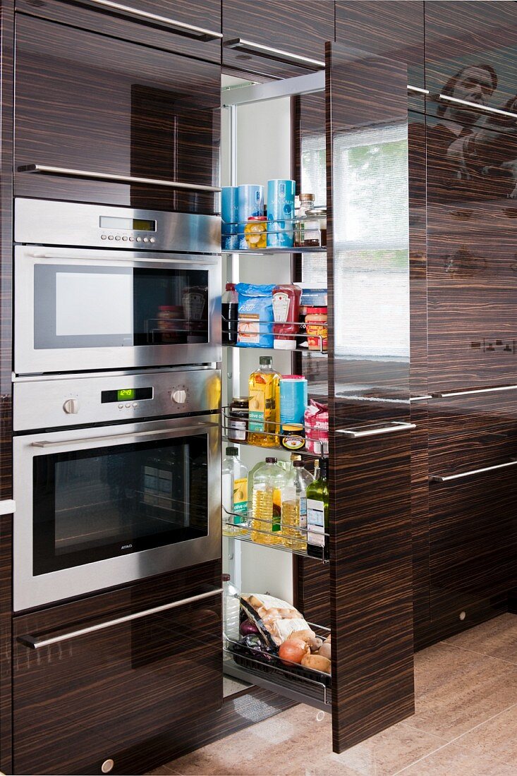 Shiny, fine wood fronts in a contemporary kitchen; a pull-out larder unit for storing groceries next to a built-in, stainless steel oven