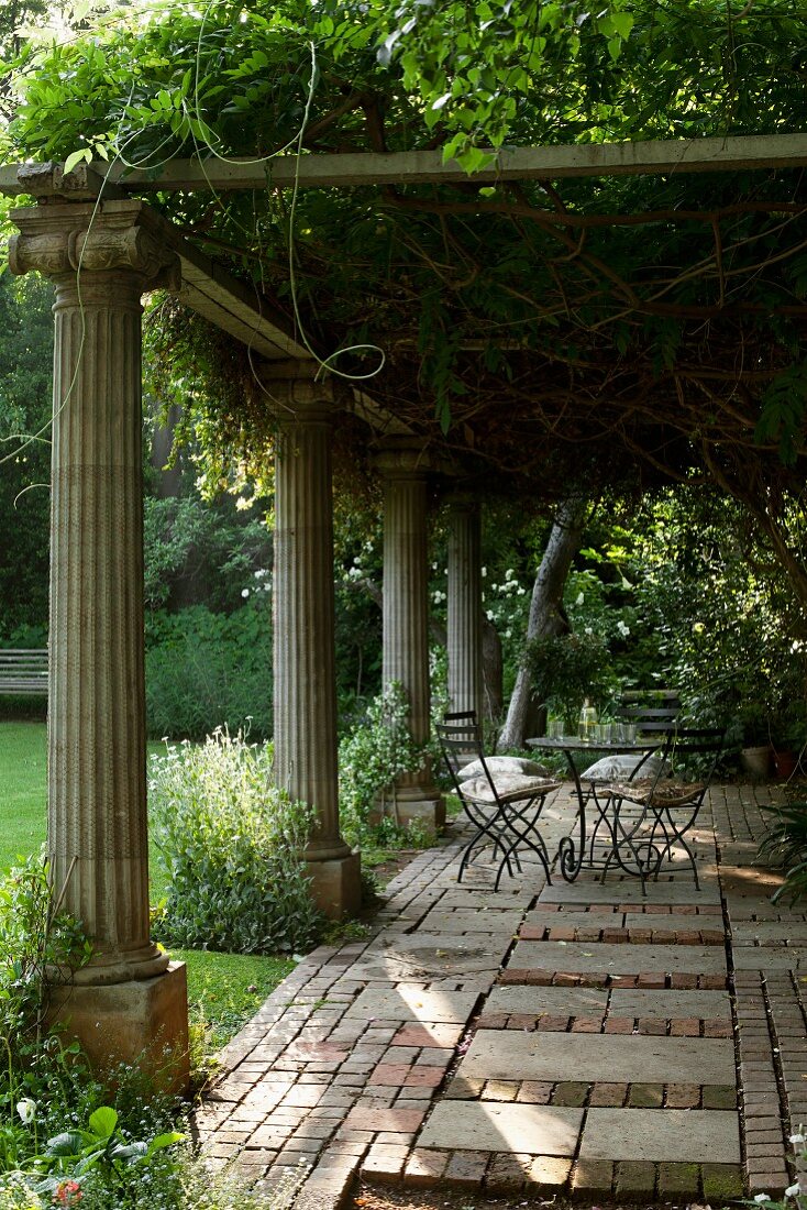 Verwunschener Sitzbereich unter berankter Pergola mit antiken Steinsäulen