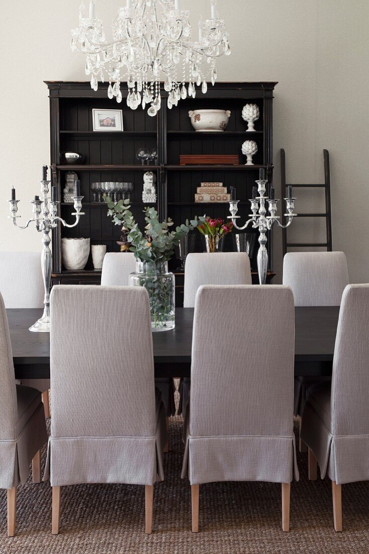 Chairs with pale grey loose covers around dining table below chandelier; antique shelves in background