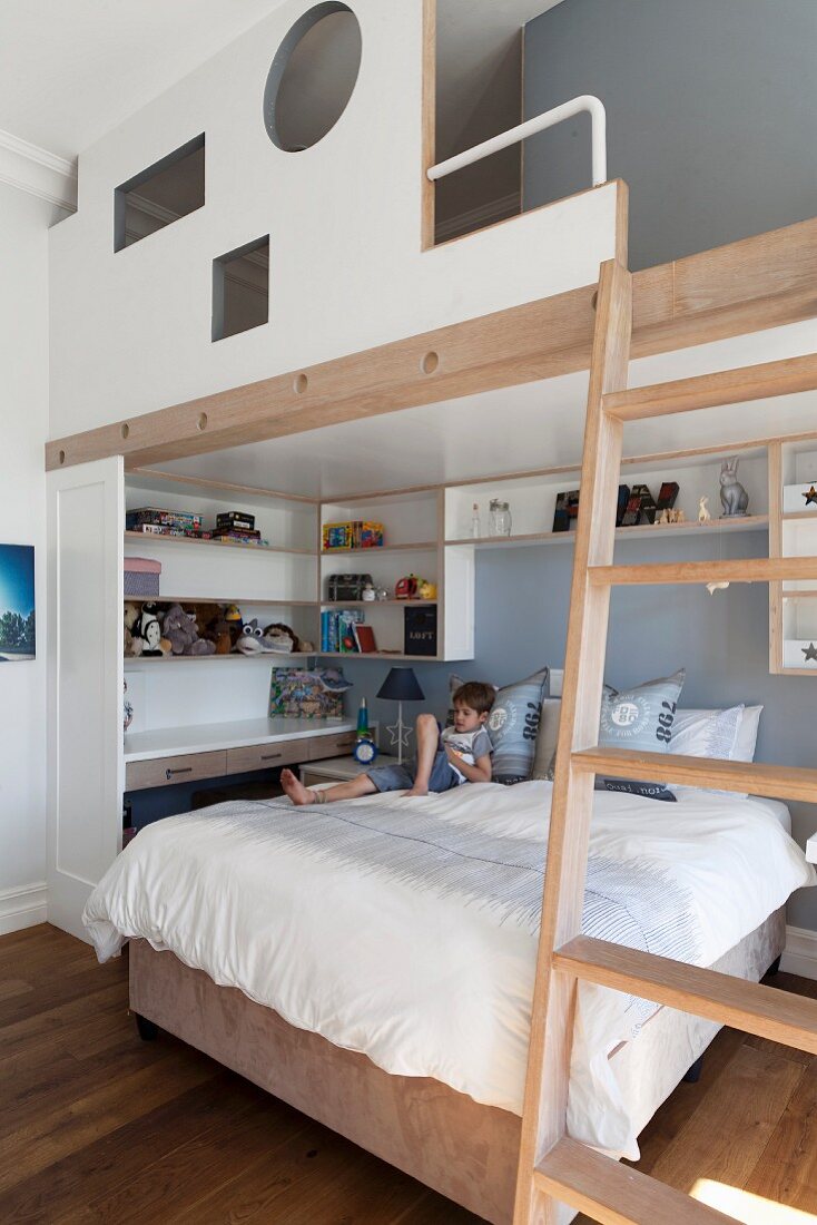 Child's bedroom with platform above bed accessed by ladder, wall-mounted shelves and little boy on bed