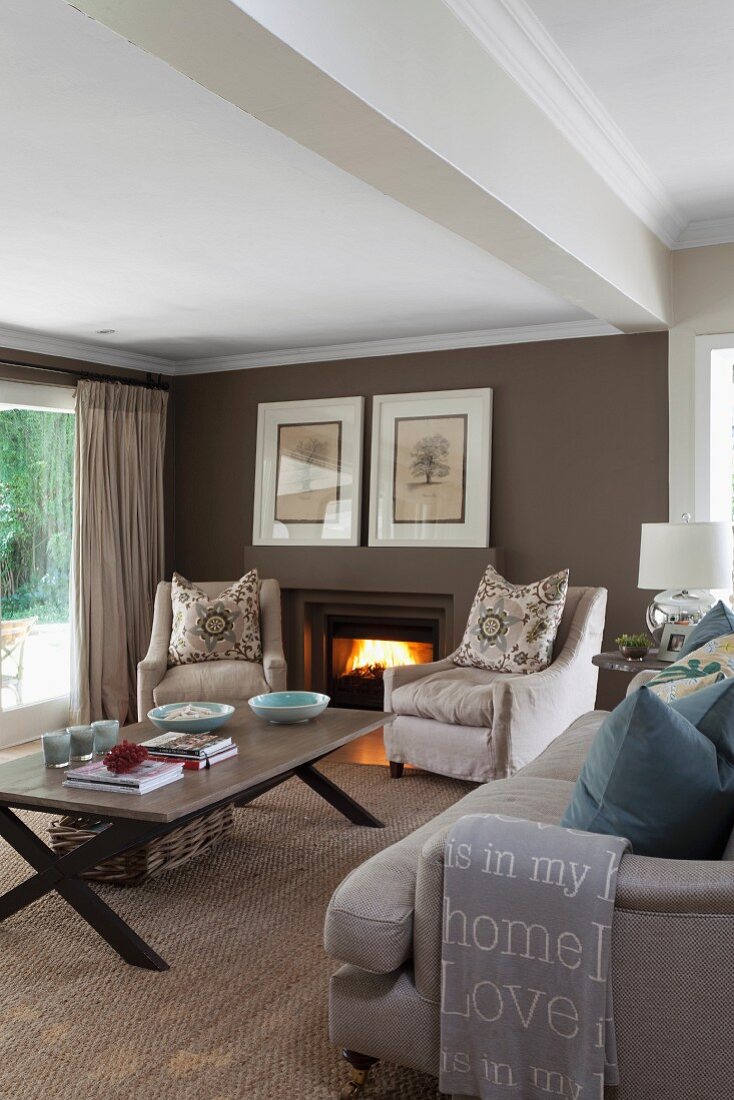 Pale armchairs with scatter cushions and sofa around coffee table on sisal carpet; open fire in brown-painted wall of living room