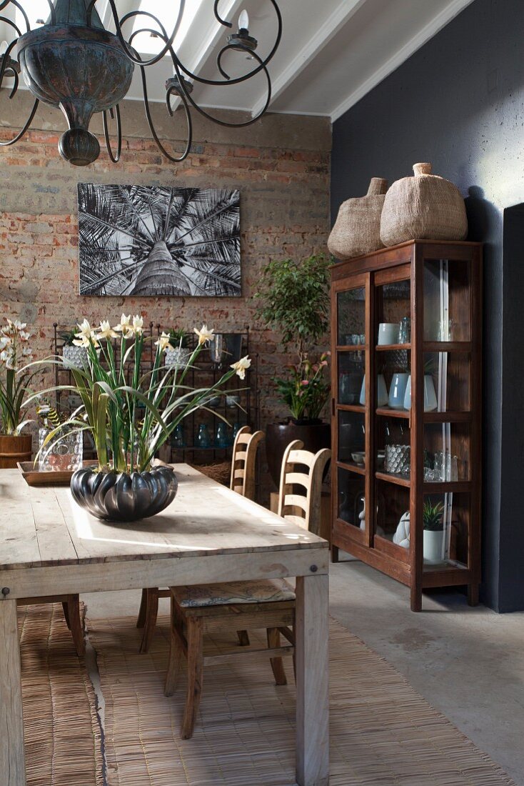 Wooden dining table and simple chairs in front of brick wall and ornate vessels in display cabinet to one side