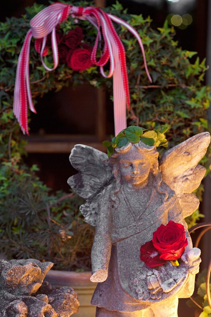 Stone angel lit from one side in front of ivy wreath in garden