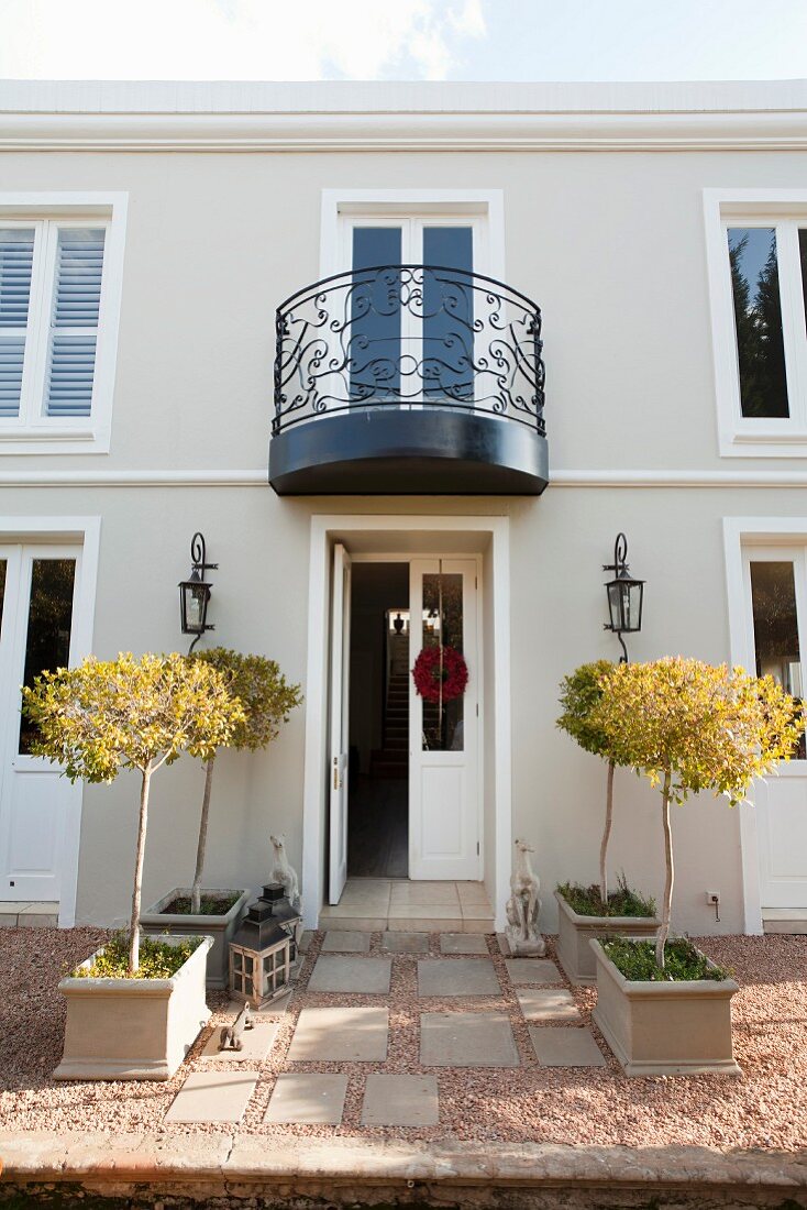 Small trees in rows of planters outside elegant house with open front door; black, wrought iron balcony on first floor