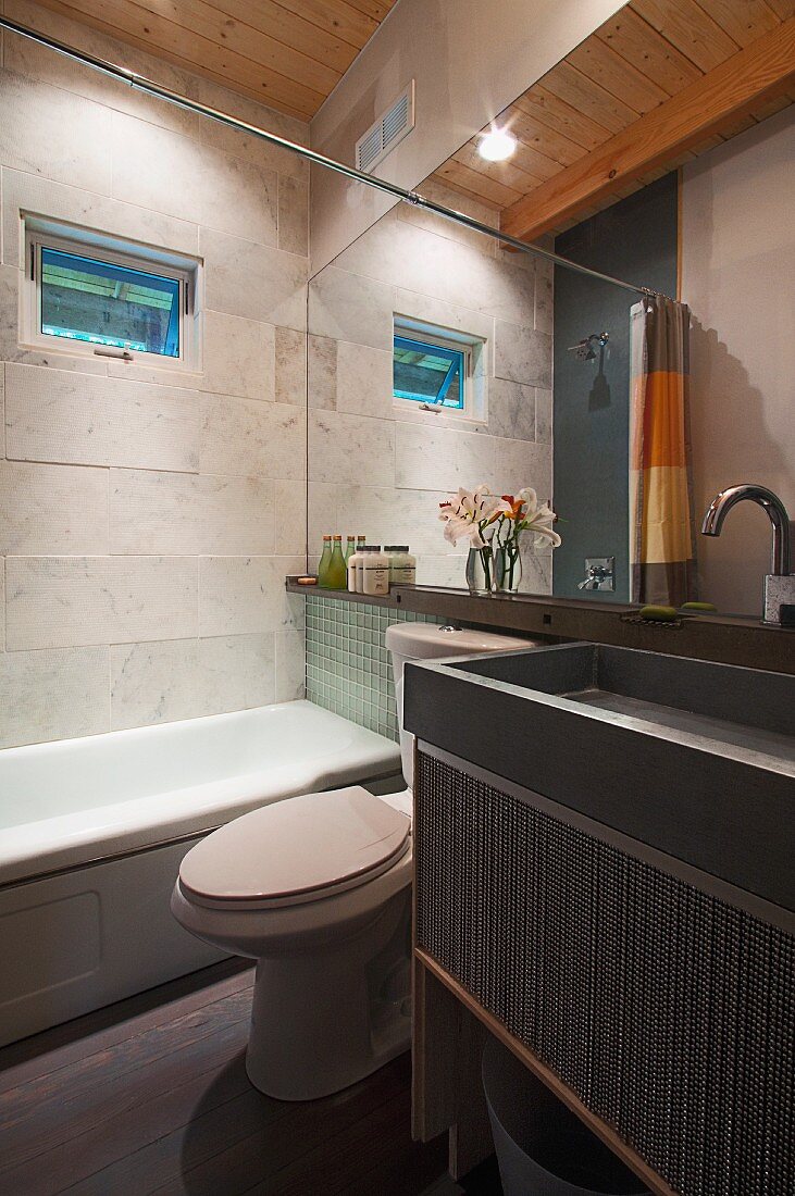 Modernes Badezimmer mit Holzdecke - trogartiges Waschbecken in Grau auf Unterbau vor Wandspiegel, daneben Badewanne an Wand mit hellgrauen Steinplatten