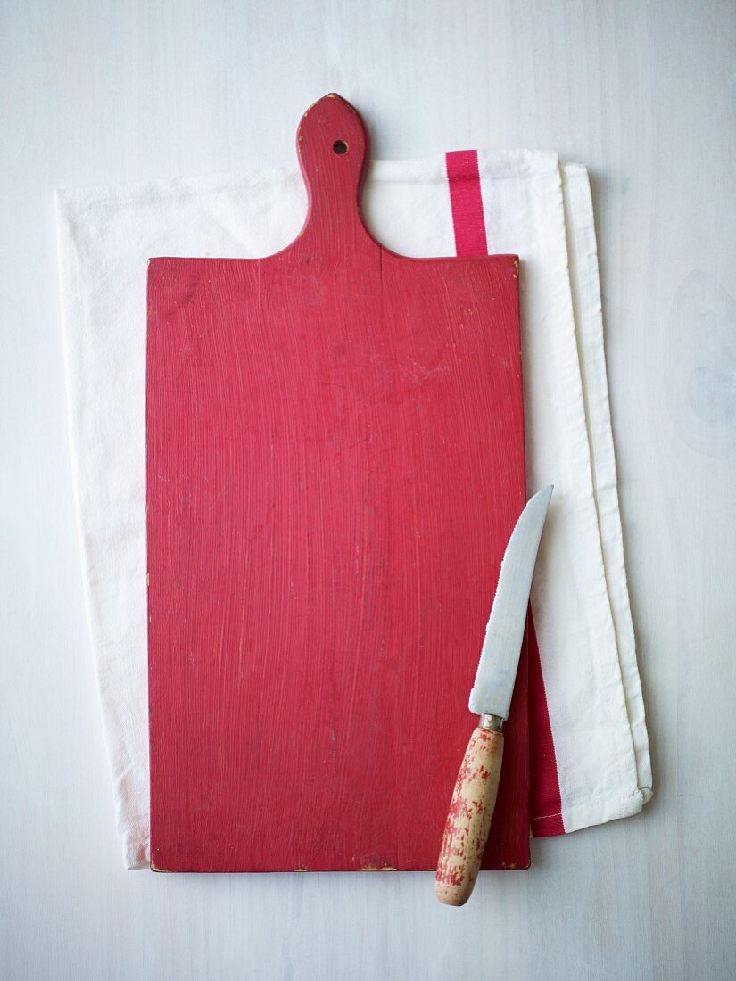 A red wooden chopping board and a knife on a tea towel