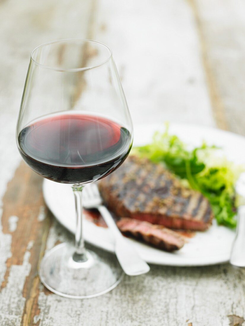 A glass of red wine in front of a grilled beef steak with salad