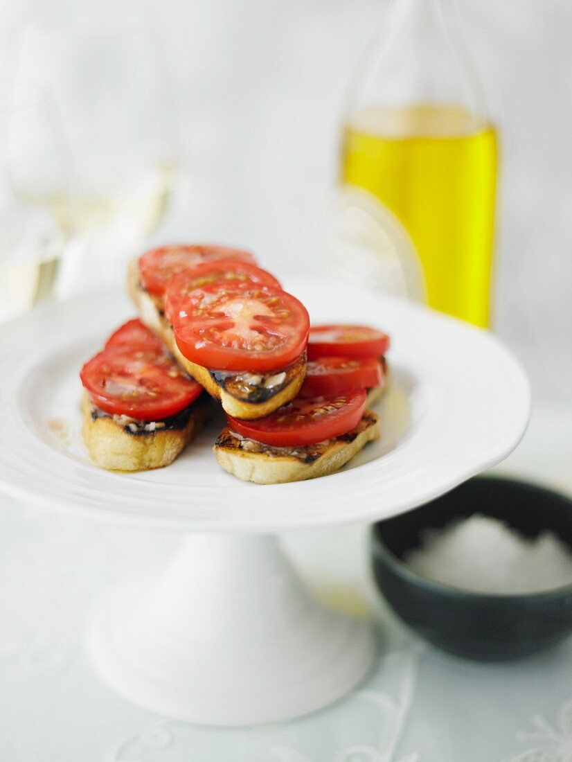 Grilled bread topped with sliced tomatoes and olive oil on a cake stand