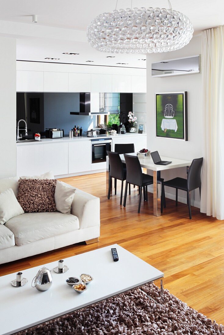 White coffee table on flokati-style rug and pale sofa in front of dining table against wall in open-plan fitted kitchen