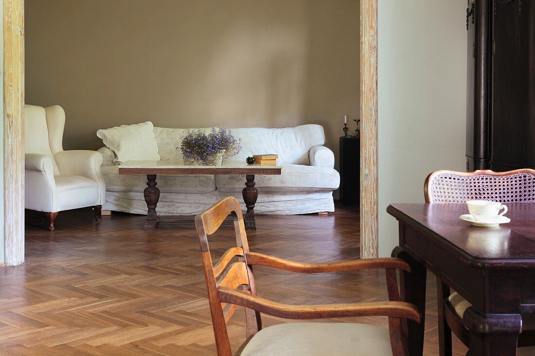 Stühle im Biedermeierstil an teilweise sichtbarem Tisch, dahinter breiter Durchgang mit Blick auf Couchtisch und weissbezogenes Sofa an grauer Wand