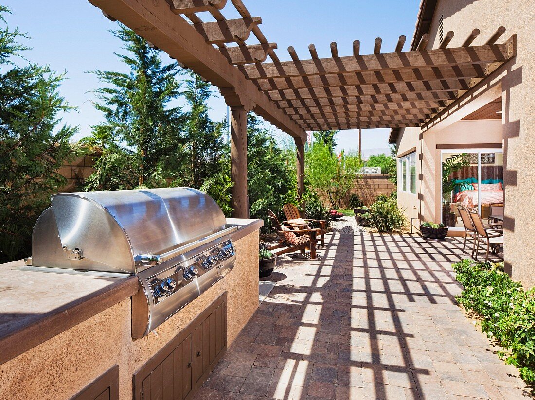 Aussengrill aus Edelstahl auf Terrasse teilweise mit Pergola aus Holz vor sonnenbeschienenem Landhaus