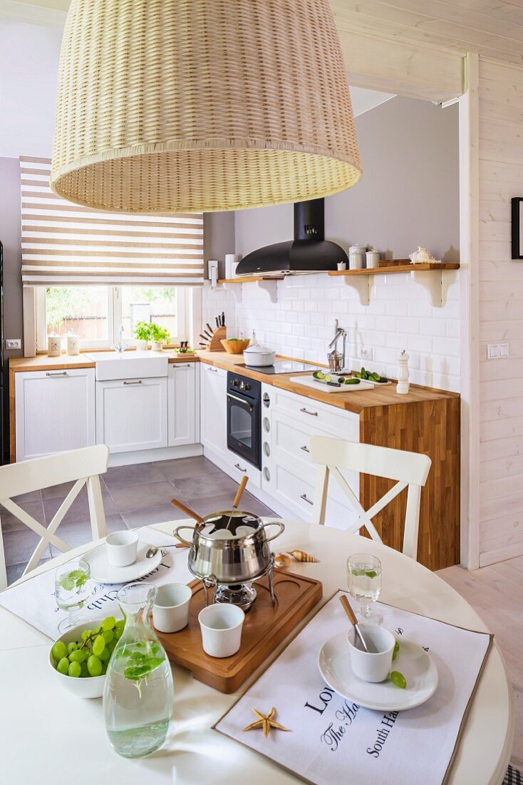 Fondue set on dining table below rattan lampshade; country-house-style fitted kitchen in background