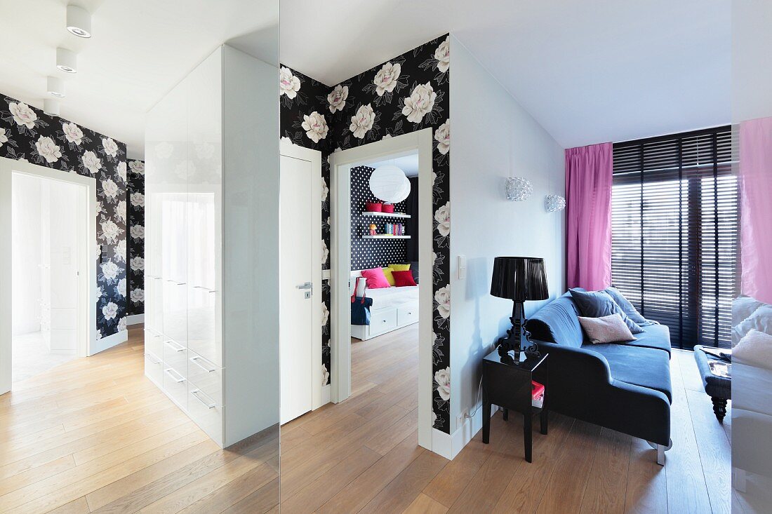 A view into a living room with a velvet sofa and a hallway with black and white floral wallpaper borders