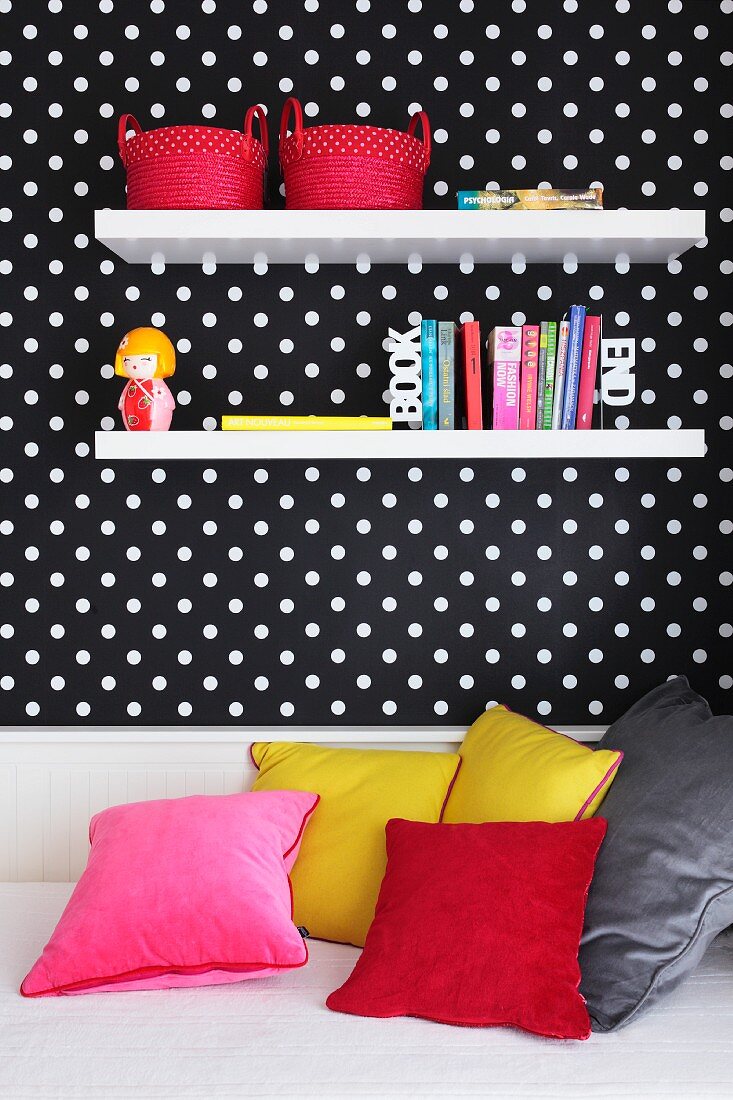 Decorative wall shelves on a wall hung with black and white spotted wallpaper above a sofa-bed with coloured cushions