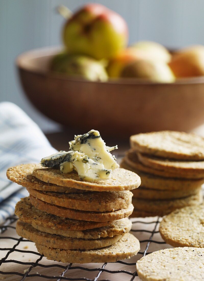A stack of poppy seed and oat biscuits with Stilton with a bowl of pears in the background