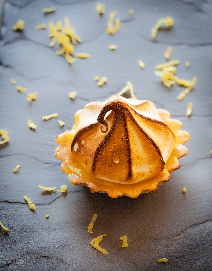 A lemon curd tartlet with a meringue topping on a grey surface surrounded by lemon zest