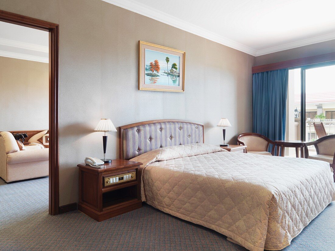 Bett mit gesteppter Tagesdecke in Hotelzimmer mit Balkon & Durchgang zum Wohnbereich