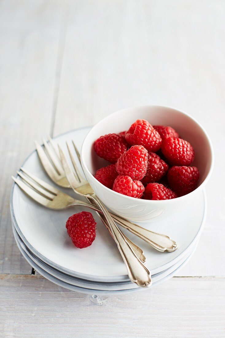 A bowl of raspberries on a stack of plates with cake forks