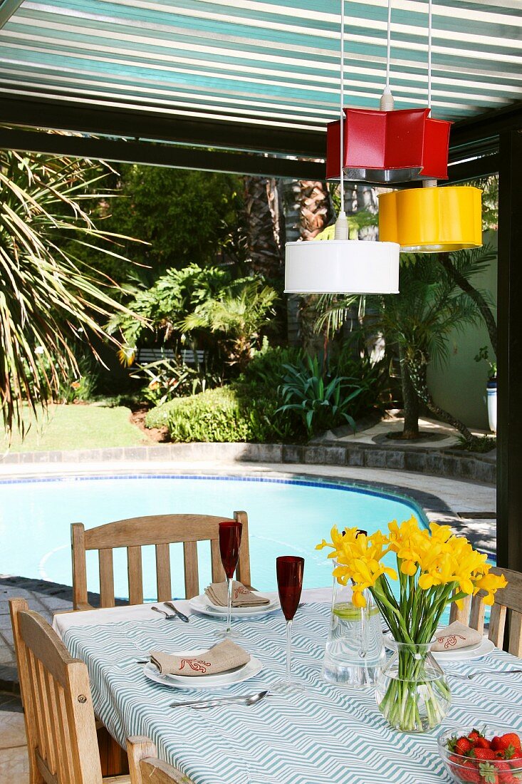 Old baking tins in different colours as pendent lamps hanging above a patio table with a pool in the background