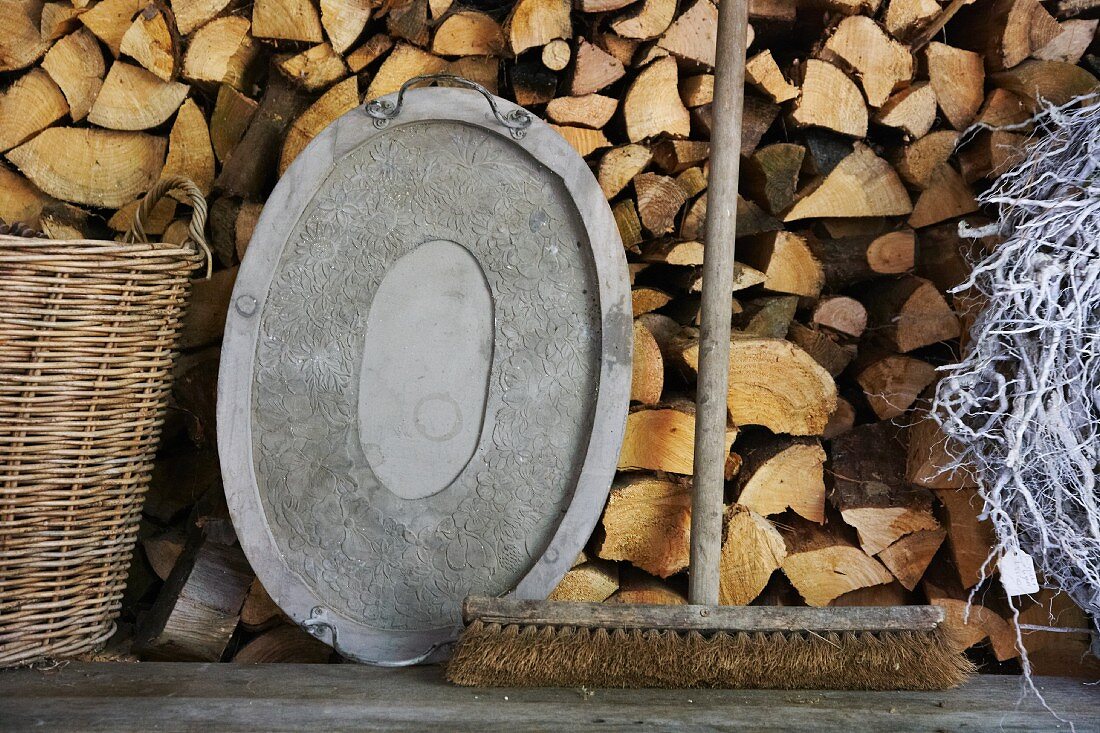An old broom and a metal tray on a wooden bench in front of a wood pile