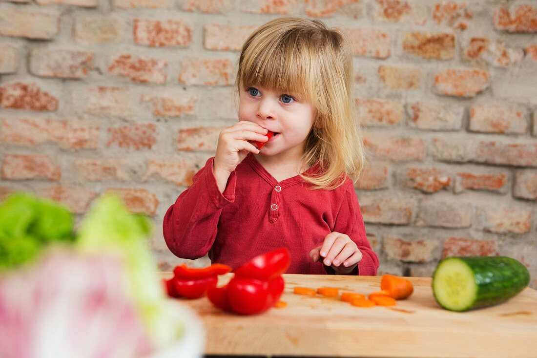 Kleines blondes Mädchen in der Küche isst rohes Gemüse