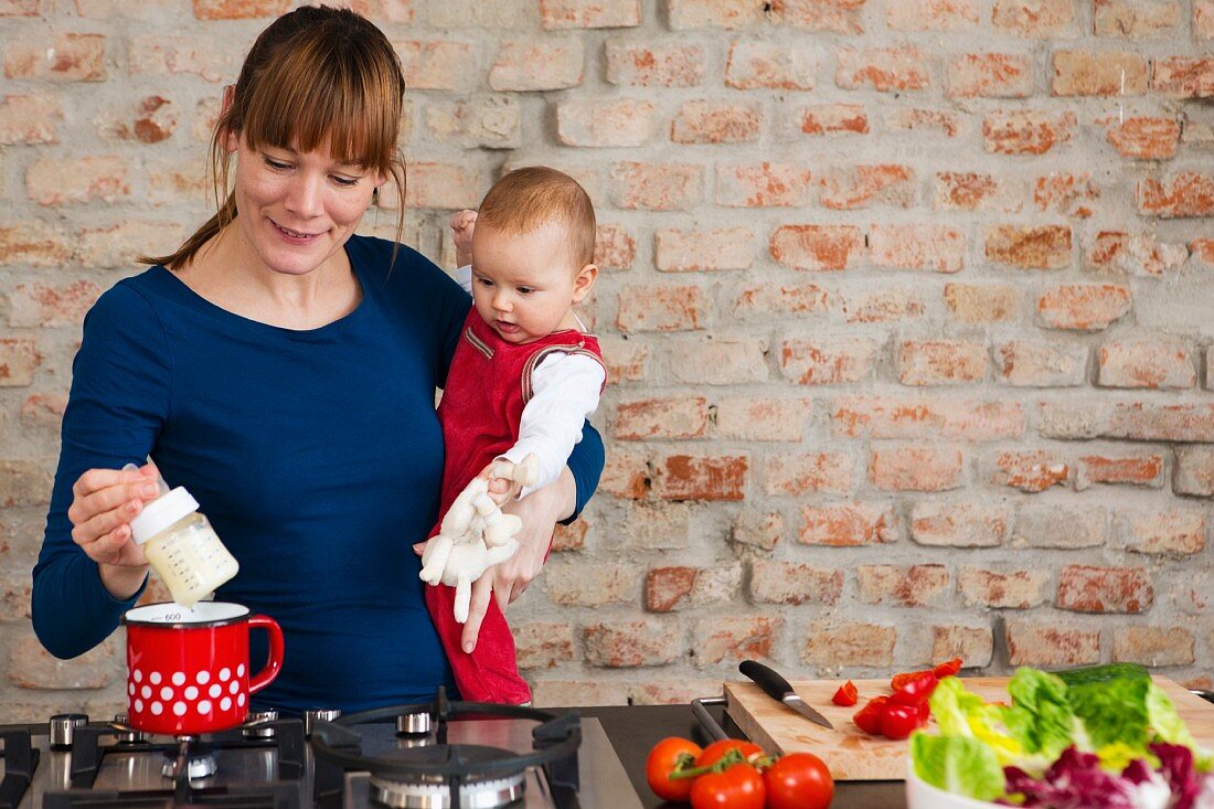 Mutter mit Baby auf dem Arm wärmt Fläschchen mit Milch in der Küche